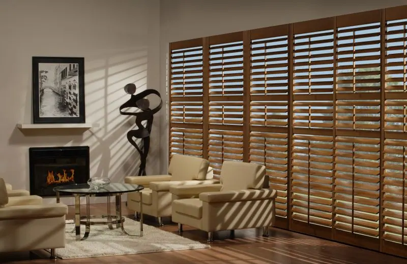 Dark wood shutters on a wide window in a living room with a fireplace