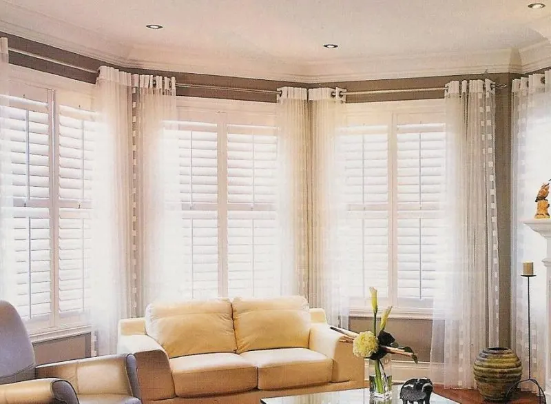 White shutters with sheer drapes in a bay window and a pale yellow sofa