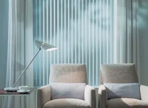Teal vertical blinds behind two grey armchairs and a reading lamp