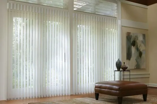 Sheer white vertical blinds on tall windows with a brown leather ottoman